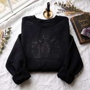 May include: A black crewneck sweatshirt featuring dark tonal embroidery of three figures standing in a circle around a central flame, surrounded by flowers, ferns, mushrooms, and a crescent moon, with the text "girls will be girls" below.