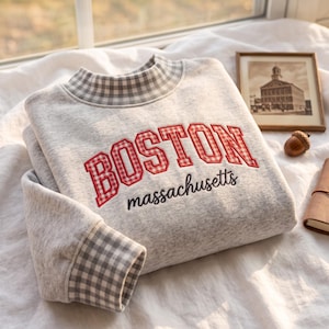 May include: A light grey heathered sweatshirt featuring a mock neck collar and sleeve cuffs with a grey and white gingham pattern. The front displays the word BOSTON in red and white gingham block letters with massachusetts in black script.