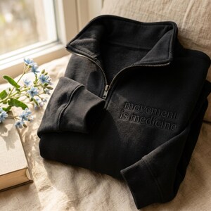 Puede incluir: Sudadera negra con cremallera hasta la mitad y la frase bordada "movement is medicine". La sudadera tiene pu&ntilde;os acanalados y cuello alto. La imagen est&aacute; tomada con luz natural, con un libro y flores en el fondo.