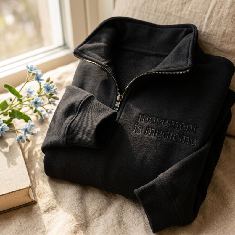 Puede incluir: Sudadera negra con cremallera hasta la mitad y la frase bordada "movement is medicine". La sudadera tiene pu&ntilde;os acanalados y cuello alto. La imagen est&aacute; tomada con luz natural, con un libro y flores en el fondo.