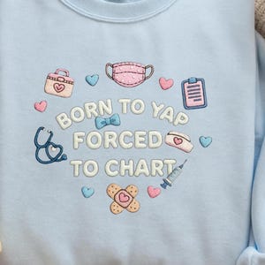 K&ouml;nnte beinhalten: Ein hellblaues Sweatshirt mit gestickten medizinischen Symbolen wie Gesichtsmaske, Stethoskop, Spritze, Pflaster und Klemmbrett. Der Text BORN TO YAP FORCED TO CHART ist in Wei&szlig; mit blauem Rand gestickt und von kleinen Herzen umgeben.