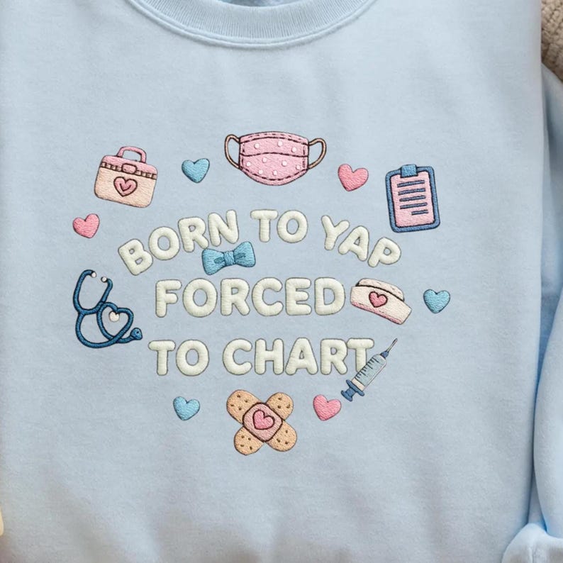 K&ouml;nnte beinhalten: Ein hellblaues Sweatshirt mit gestickten medizinischen Symbolen wie Gesichtsmaske, Stethoskop, Spritze, Pflaster und Klemmbrett. Der Text BORN TO YAP FORCED TO CHART ist in Wei&szlig; mit blauem Rand gestickt und von kleinen Herzen umgeben.