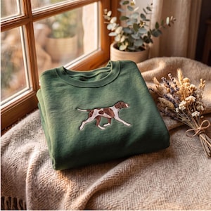 May include: A forest green crewneck sweatshirt featuring an embroidered graphic of a brown and white spotted dog in a running pose. The sweatshirt has a ribbed collar and long sleeves, with the dog design centered on the chest area of the garment.