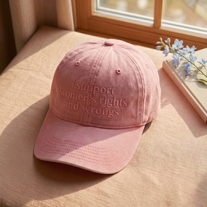 Op de afbeelding: Een roze baseballpet met de geborduurde tekst "i support women's rights and wrongs". De pet heeft een gebogen klep en een ronde kroon. De hoed is gemaakt van een zachte, getextureerde stof.