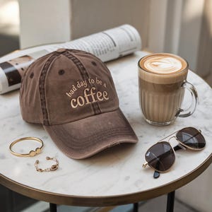 May include: A brown baseball cap with the embroidered text "bad day to be a coffee" sits on a marble table next to a latte in a clear glass mug and a pair of sunglasses. A gold bracelet and a newspaper are also on the table.
