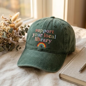 May include: A green baseball cap with the embroidered text "support your local library" in colorful thread. A rainbow is embroidered below the text. The hat is sitting on a surface near a window.