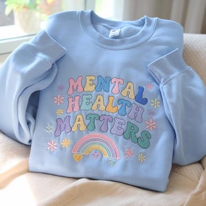May include: A light blue sweatshirt with the embroidered text "MENTAL HEALTH MATTERS" in colorful, retro-style lettering. The design includes a rainbow and floral accents. The sweatshirt is folded on a neutral surface.