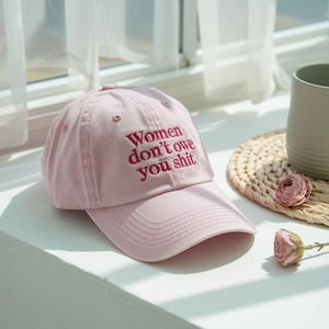 Könnte beinhalten: Hellrosa Baseballkappe mit gebogenem Schirm. Auf der Kappe ist in dunklerem Rosa der Text "Women don't owe you shit" gestickt. Die Kappe liegt auf einer weißen Oberfläche.