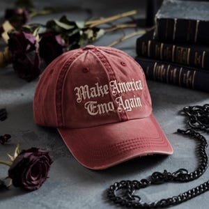 May include: A distressed burgundy baseball cap with the phrase "Make America Emo Again" embroidered in white gothic lettering. The cap has a curved brim and a worn appearance, suggesting a vintage or retro style. The background includes dark roses and books.