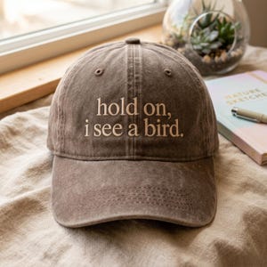 Może przedstawiać: Brązowa czapka bejsbolówka z wyhaftowanym napisem "hold on, i see a bird." Czapka ma zakrzywiony daszek i jest wykonana z miękkiego, zużytego materiału. Kapelusz leży na beżowej powierzchni.