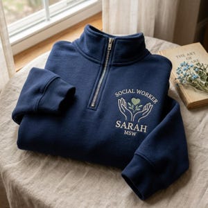 Op de afbeelding: Marineblauwe sweatshirt met een kwartrits en de witte geborduurde tekst "SOCIAL WORKER SARAH MSW". Het ontwerp bevat een hart en bladeren. De sweatshirt is opgevouwen op een tafel.