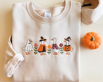 May include: Beige sweatshirt with embroidered Halloween geese. The geese are dressed in costumes: a ghost, a witch, a vampire, and a jack-o'-lantern. A matching neck warmer and a small orange pumpkin are also in the image.