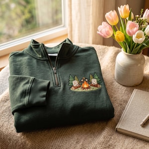 Könnte beinhalten: Ein dunkelgrünes Sweatshirt mit kurzem Reißverschluss und einer Stickerei auf der Brust. Das Motiv zeigt zwei lächelnde Marshmallows, die auf Baumstämmen an einem Lagerfeuer sitzen, umgeben von Tannen. Das Sweatshirt hat einen Stehkragen.