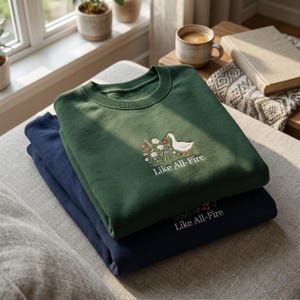 May include: A dark green crewneck sweatshirt stacked on top of a navy blue sweatshirt. The green sweatshirt features an embroidered design of a white goose standing among colourful wildflowers with the text Like All-Fire stitched below it.