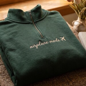 Puede incluir: Una sudadera verde oscuro con cuello alzado y cremallera de un cuarto. La frase "airplane mode" est&aacute; bordada en cursiva, acompa&ntilde;ada de un peque&ntilde;o gr&aacute;fico de avi&oacute;n. La sudadera est&aacute; doblada sobre una superficie texturizada.