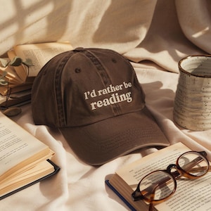 May include: A brown baseball cap with the text "I'd rather be reading" in white. The cap is sitting on a white surface next to open books, a ceramic mug, and eyeglasses.