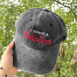 May include: A dark grey baseball cap featuring the embroidered text "i need a Coke Zero" in red. The cap has a curved peak and a traditional six-panel construction, suitable for everyday use.