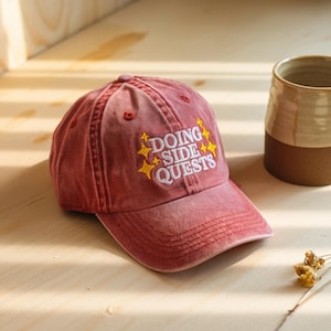 Puede incluir: Gorra de b&eacute;isbol roja con la frase "DOING SIDE QUESTS" bordada en amarillo y blanco. La gorra tiene una visera curva y un aspecto desgastado. En el fondo se ve una taza de cer&aacute;mica.