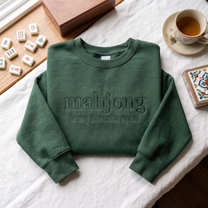 Może przedstawiać: Leśnozielona bluza z okrągłym dekoltem i wyhaftowanym napisem "mahjong is my favorite sport" pasującą ciemnozieloną nicią. Odzież posiada długie rękawy, prążkowany dekolt oraz prążkowane mankiety, zaprezentowana w płaskim układzie.