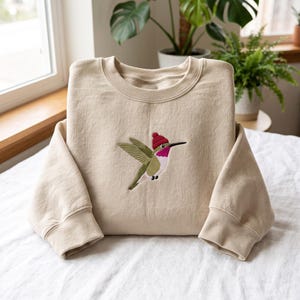 May include: A beige crewneck sweatshirt featuring an embroidered graphic of a hummingbird in flight. The bird has olive green wings, a white chest, a magenta throat patch, and is wearing a small, solid red knit beanie hat on its head.