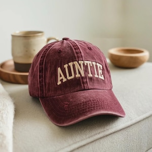 Könnte beinhalten: Burgunderfarbene Baseballkappe mit dem Wort "AUNTIE" in cremefarbenen Blockbuchstaben gestickt. Die Kappe hat einen Used-Look und einen gebogenen Schirm. Ein lässiges Accessoire.
