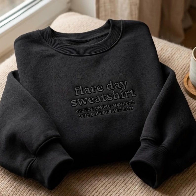 Puede incluir: Sudadera negra con el texto "flare day sweatshirt" y la frase "caution please approach with patience and love" en un tono a juego. La sudadera tiene cuello redondo y mangas largas.