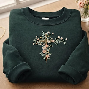 Pode incluir: Camisola verde escuro com um design de cruz floral bordada. A cruz é adornada com flores rosa e amarelas e folhas verdes. A camisola tem gola redonda e mangas compridas.