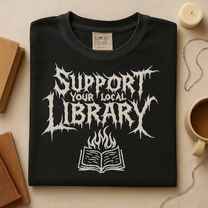 Puede incluir: Camiseta negra de cuello redondo con el texto "SUPPORT YOUR LOCAL LIBRARY" en una fuente gótica. Debajo del texto hay una ilustración de un libro abierto con llamas. La camiseta tiene una etiqueta Comfort Colors.