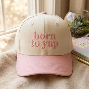 May include: A cream-colored baseball cap with a pink brim and a pink embroidered text on the front that reads "born to yap". The cap features a structured crown with a small button at the top and stitched eyelets for ventilation.