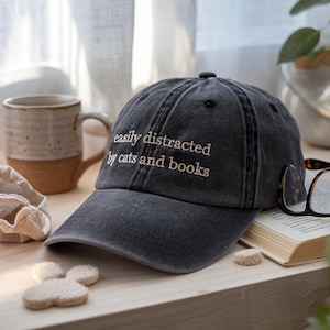 May include: A dark gray baseball cap with the embroidered text "easily distracted by cats and books." The cap is sitting on a wooden surface next to a coffee mug, a book, and a pair of glasses.
