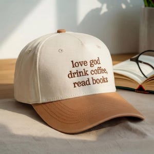 May include: A cream-colored baseball cap with a tan brim and a small tan button on top. The front of the cap features brown embroidered text that reads, love god drink coffee, read books. The cap has a structured crown and stitched brim details.
