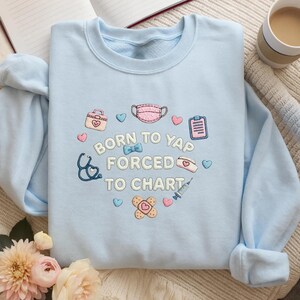 K&ouml;nnte beinhalten: Ein hellblaues Sweatshirt mit Rundhalsausschnitt und gestickten medizinischen Motiven wie Stethoskop, Gesichtsmaske, Spritze, Pflaster und Klemmbrett sowie dem wei&szlig;en Schriftzug "BORN TO YAP FORCED TO CHART", umgeben von kleinen Herzen.