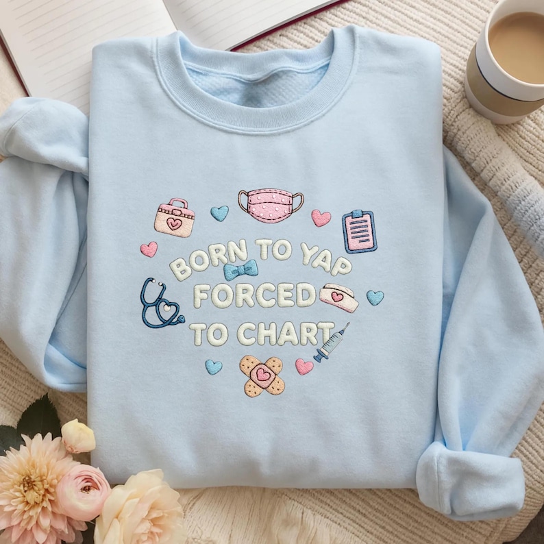 K&ouml;nnte beinhalten: Ein hellblaues Sweatshirt mit Rundhalsausschnitt und gestickten medizinischen Motiven wie Stethoskop, Gesichtsmaske, Spritze, Pflaster und Klemmbrett sowie dem wei&szlig;en Schriftzug "BORN TO YAP FORCED TO CHART", umgeben von kleinen Herzen.