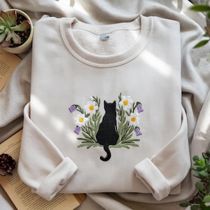 May include: Cream-colored sweatshirt with an embroidered design of a black cat silhouette surrounded by white daisies, purple bellflowers, and green leaves. The sweatshirt has a crew neck and cuffed sleeves.
