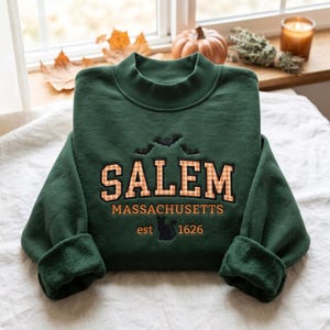 Könnte beinhalten: Ein dunkelgrünes Langarm-Sweatshirt mit Stehkragen. Auf der Vorderseite befinden sich gestickte schwarze Fledermäuse über dem Schriftzug SALEM in orange-weißem Vichy-Muster, gefolgt von MASSACHUSETTS, einer schwarzen Katzensilhouette und est 1626 in Orange.