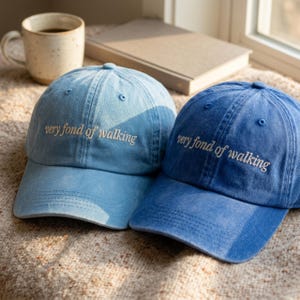 May include: Two baseball caps in light blue and medium blue denim fabric. Each cap features the embroidered text "very fond of walking" in a light-coloured thread across the front panel. The caps have a curved brim and a six-panel construction.