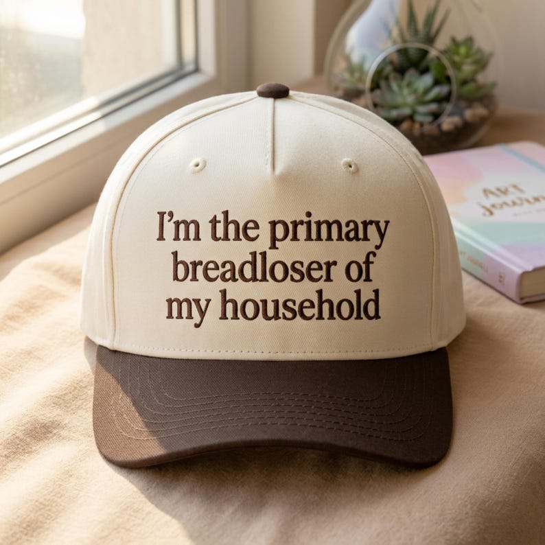 K&ouml;nnte beinhalten: Baseballcap mit cremefarbener Krone und dunkelbraunem Schirm. Der Aufdruck lautet "I'm the primary breadloser of my household" in dunkelbraunen Buchstaben. Oben befindet sich ein brauner Knopf.