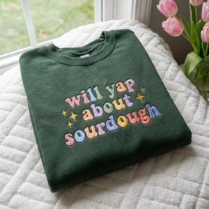 Può includere: Una felpa verde scuro con la scritta ricamata "will yap about sourdough" in colori pastello. Il testo è ad arco e include accenti di stelle. La felpa è piegata su una superficie bianca e strutturata.