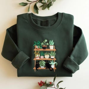 Peut inclure: Un sweat-shirt à col rond vert forêt foncé avec une broderie représentant une étagère en bois à trois niveaux. L'étagère contient diverses plantes en pot et deux chats noirs assis sur le niveau inférieur parmi le feuillage vert et blanc.