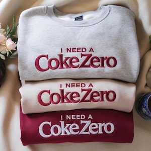 May include: Three sweatshirts in gray, cream, and burgundy, stacked on top of each other. Each sweatshirt has the embroidered text "I NEED A Coke Zero" in red. The sweatshirts are folded and arranged neatly.
