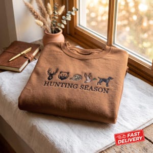 Könnte beinhalten: Ein rostfarbenes Sweatshirt mit Rundhalsausschnitt und gestickten Symbolen eines Hirschkopfes, eines Bärengesichts, einer Tarnkappe, einer fliegenden Ente und eines Hundes, mit dem Text "HUNTING SEASON" in schwarzem Garn unter den Motiven.