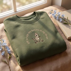 May include: An olive green crew neck sweatshirt. The design features an embroidered raccoon on a scooter, wearing a hat, with the text "Off to help tiny humans do big things" in a curved arc around the raccoon.