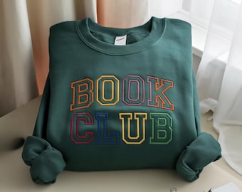 Puede incluir: Una sudadera verde oscuro con la inscripción bordada "BOOK CLUB". Las letras son multicolores, con contornos en naranja, amarillo, rosa, azul y verde. La sudadera está doblada sobre una superficie clara.