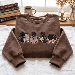 Pode incluir: Uma camisola castanha de gola redonda com um bordado de quatro pequenos dragões pretos. Um dragão empilha livros, um está sob uma manta estampada, um usa óculos enquanto lê e um dorme numa almofada ao lado de um tablet e uma chávena.