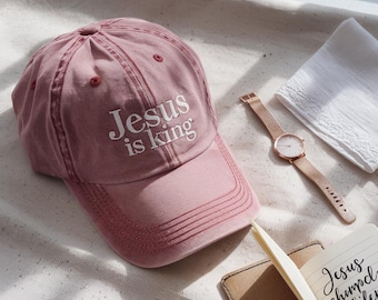 May include: A dusty rose baseball cap with the text "Jesus is king" embroidered in white. A rose gold watch, a white folded cloth, and a small notebook are also visible.