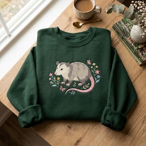 May include: A dark forest green crewneck sweatshirt featuring a central embroidered graphic of a grey opossum surrounded by colourful small flowers, green leaves, and tiny butterflies. The opossum has a pink nose, pink ears, and a long pink tail.