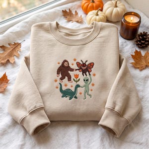 May include: A beige crewneck sweatshirt featuring an embroidered design of a brown Bigfoot, a moth with orange-spotted wings, a green alien, and a blue sea creature holding hands around a central orange tulip, accented by small orange star shapes.
