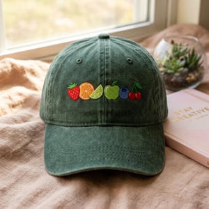 May include: A dark green baseball cap with embroidered fruit: a strawberry, orange slice, lime, green apple, blueberry, and cherries. The cap is made of a soft fabric and has a curved brim. The cap is sitting on a light brown surface.