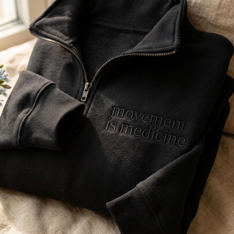 Puede incluir: Sudadera negra con cierre de cremallera de cuarto y pu&ntilde;os acanalados. El texto "movement is medicine" est&aacute; bordado en la parte delantera. La sudadera tiene un cuello alto y un ajuste relajado.