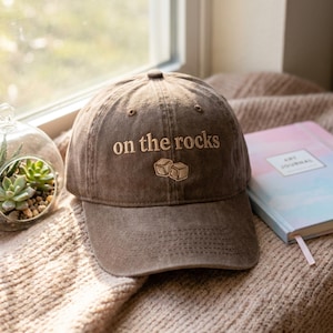 May include: A brown baseball cap with the embroidered text "on the rocks" and two ice cube graphics. The cap is on a textured surface next to a terrarium and a notebook.